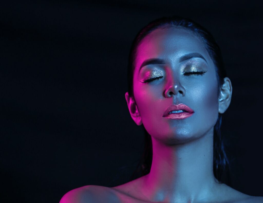 En fortryllende kvinne med glitrende sminke, venstre side av ansiktet badet i fuchsia lys, gulløyelokk, mot en mystisk mørk blå bakgrunn